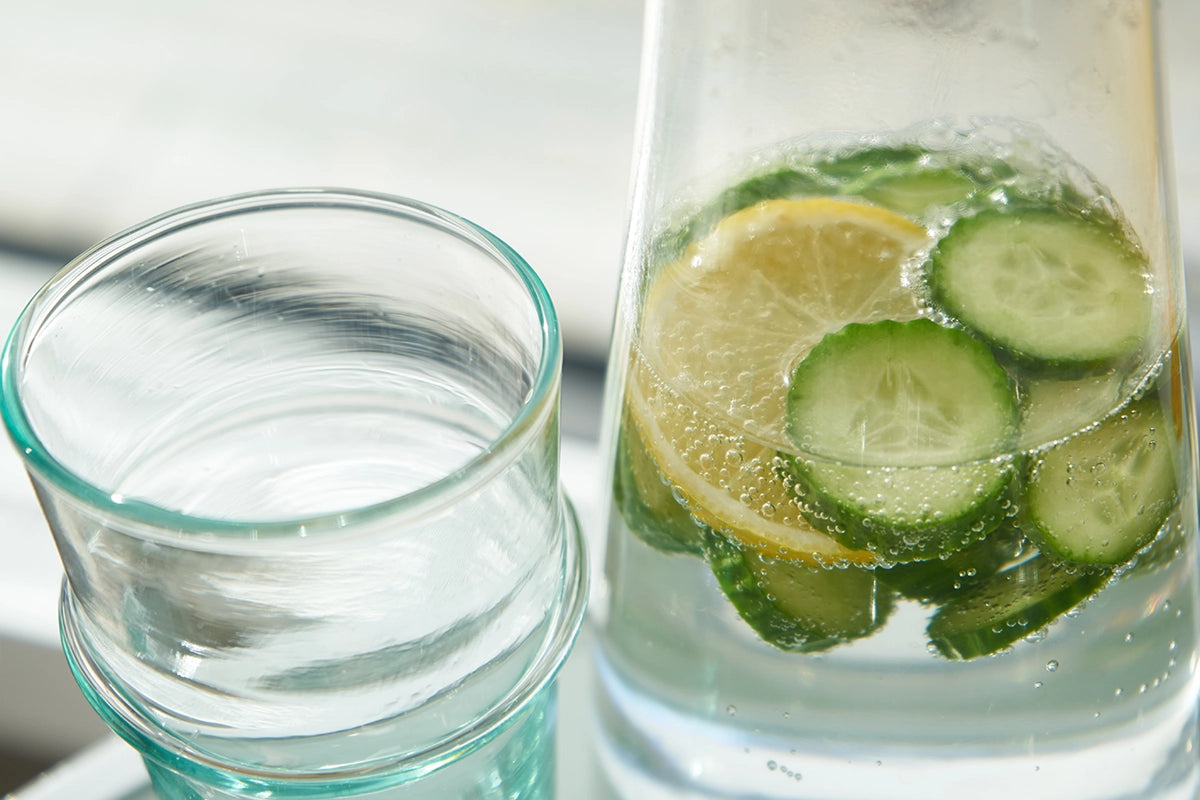 Wasserkaraffe mit Gurkenscheiben und ein Glas Wasser als Anregung, über den Tag verteilt regelmäßig zu trinken.