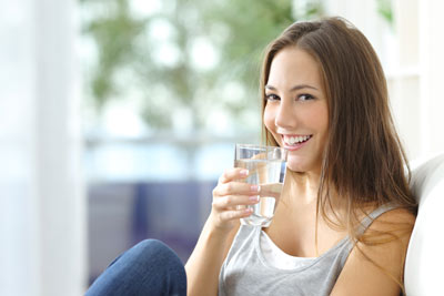 Frau trinkt ein Glas Wasser: Fazit der Studienlage ist eine tägliche Trinkmenge von 1.410 Millilitern, für Falten zählt vor allem Sonnenschutz.